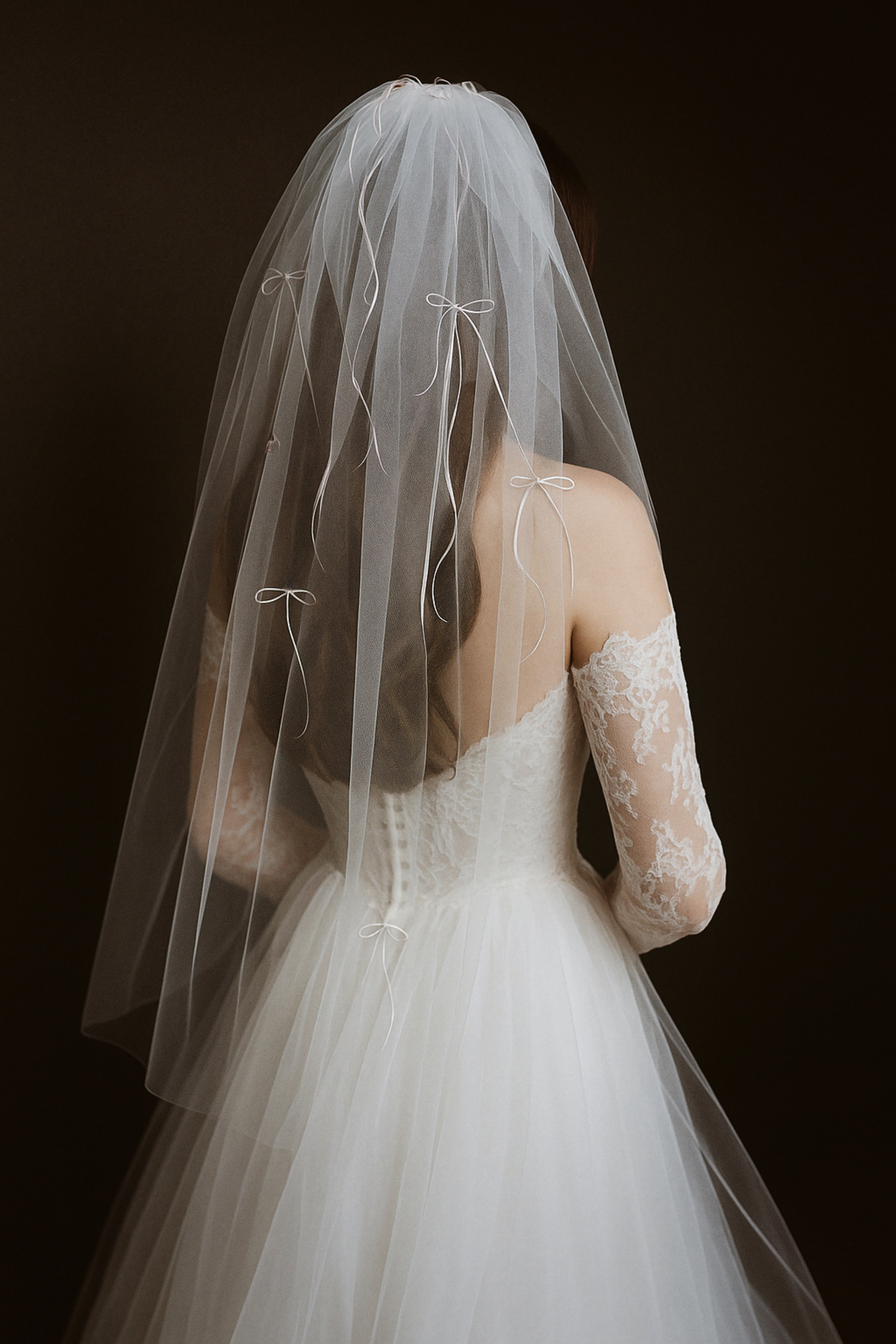 A bride wears a fingertip length wedding veil with mini satin bows in shades of white.