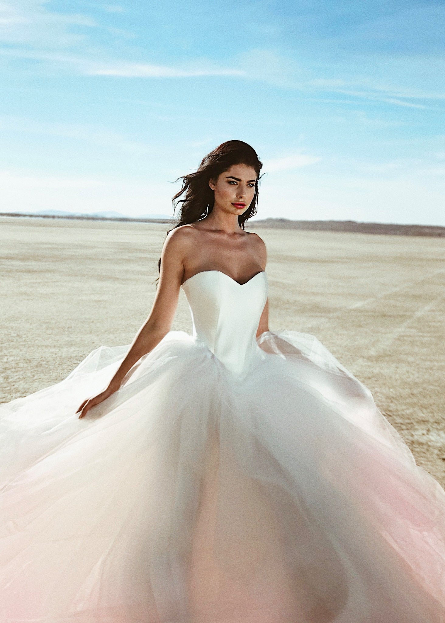 A model wears a satin Basque waist wedding dress with blush ombre tulle skirt on a dry lake bed.