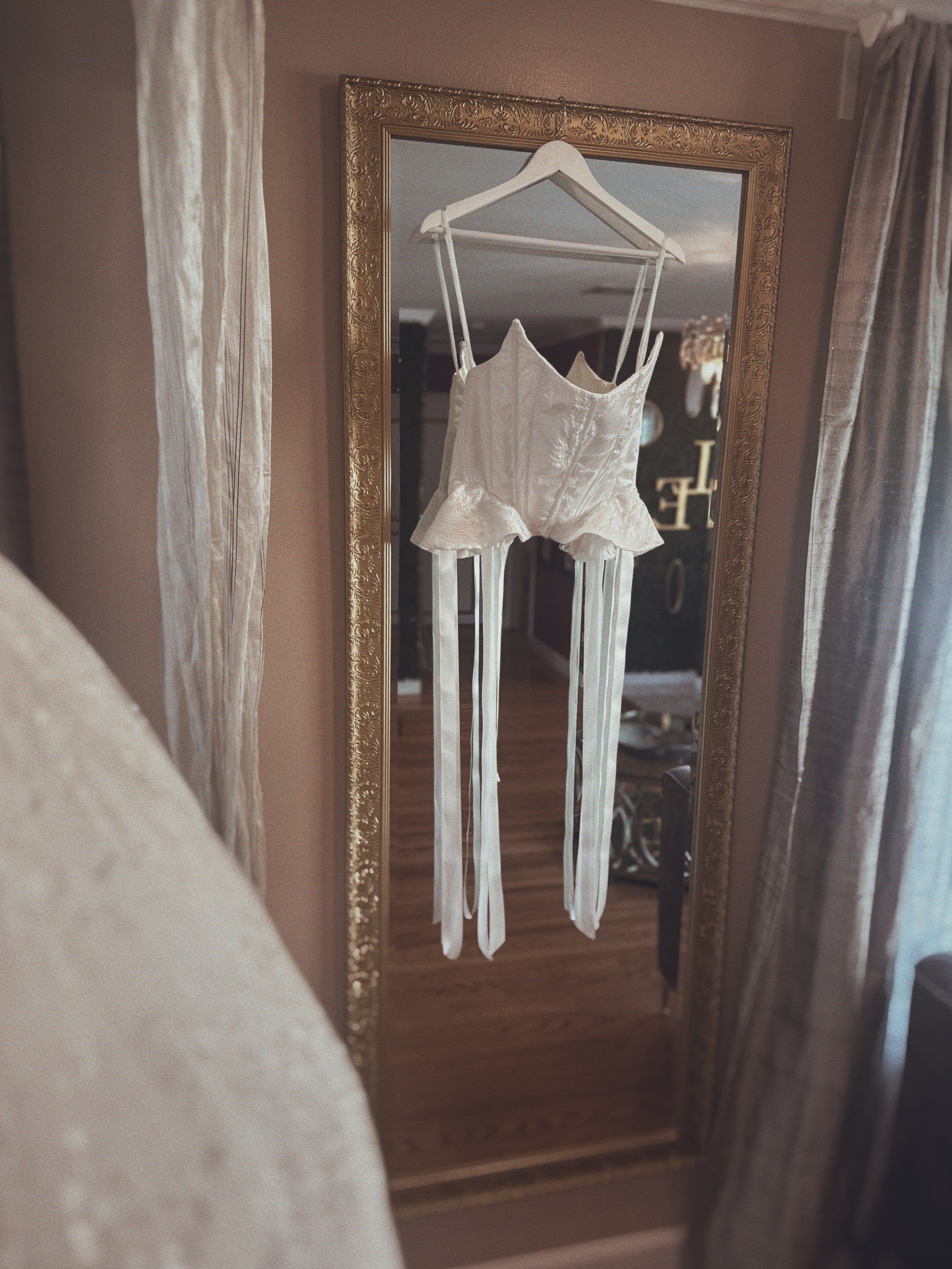 White bridal corset top with basque waist and peplum detail, shown hanging in a vintage mirror
