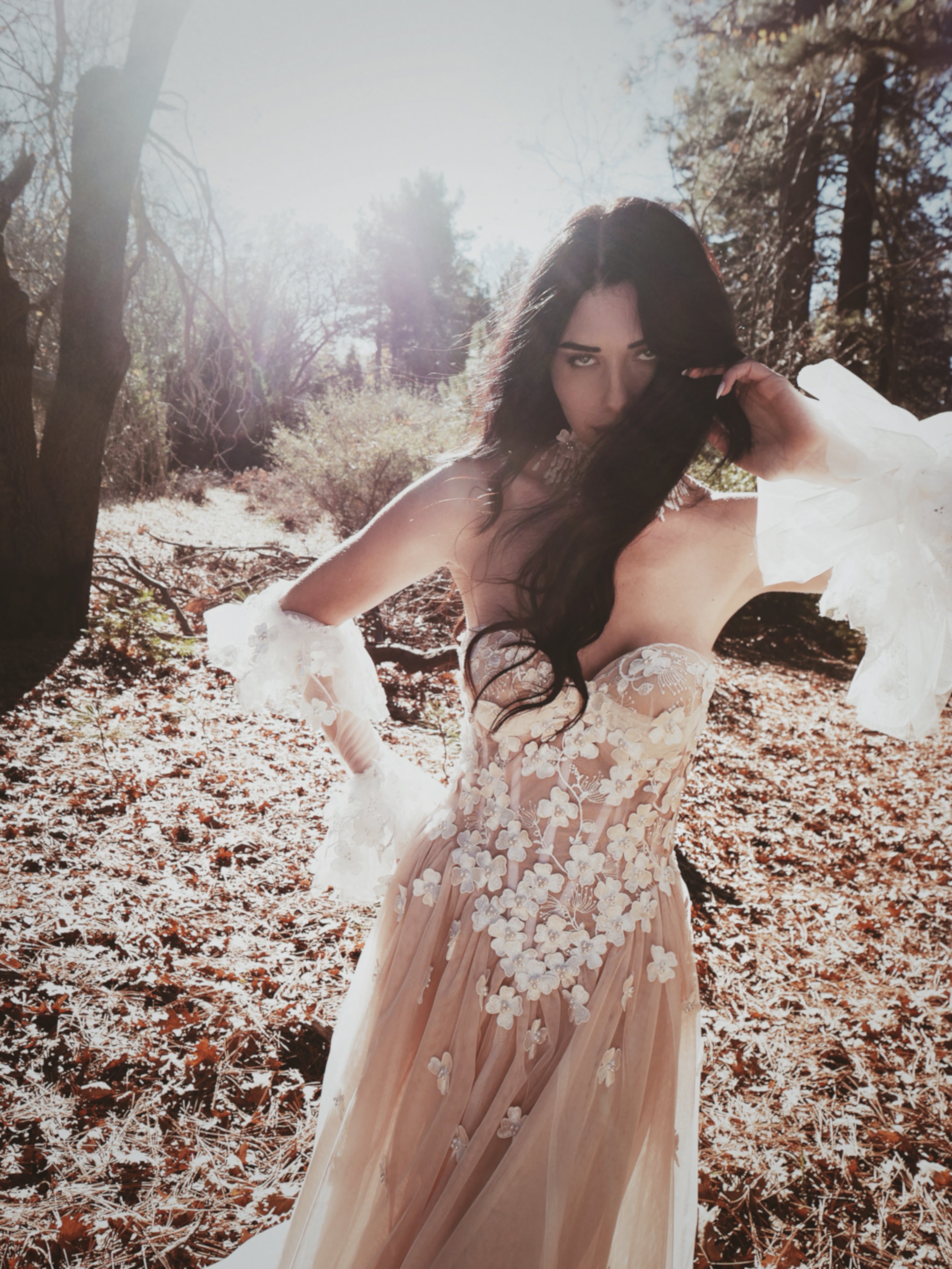 Bride in a floral wedding dress standing in a forest with sunlight filtering through the trees.