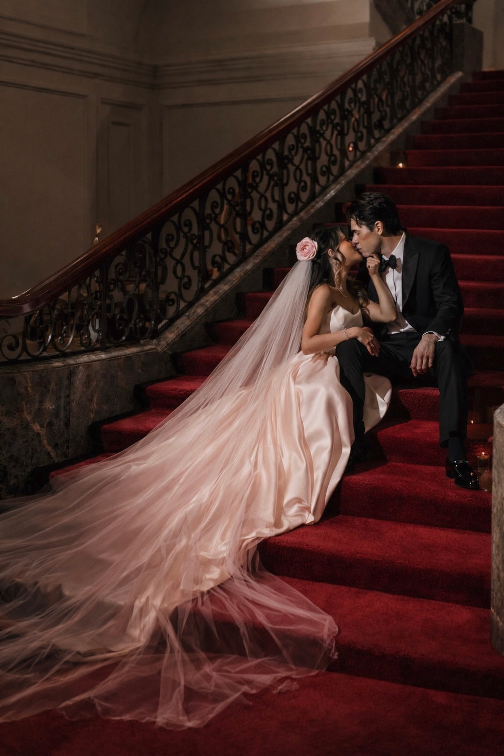 A bride wears a blush pink silk taffeta rose wedding veil on a red velvet staircase with groom.