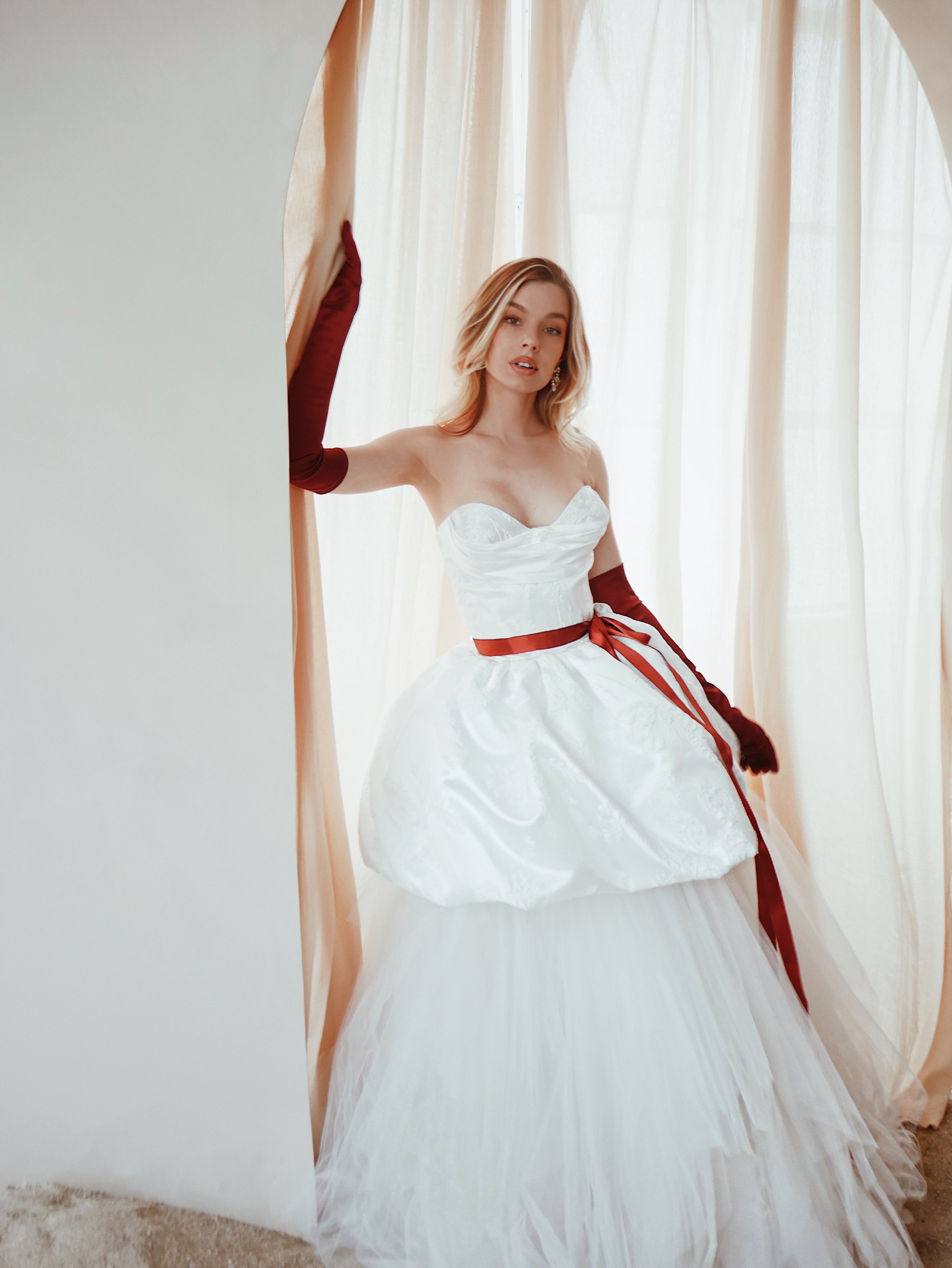 A model poses in a satin corset wedding dress with bubble hem and red satin opera gloves.
