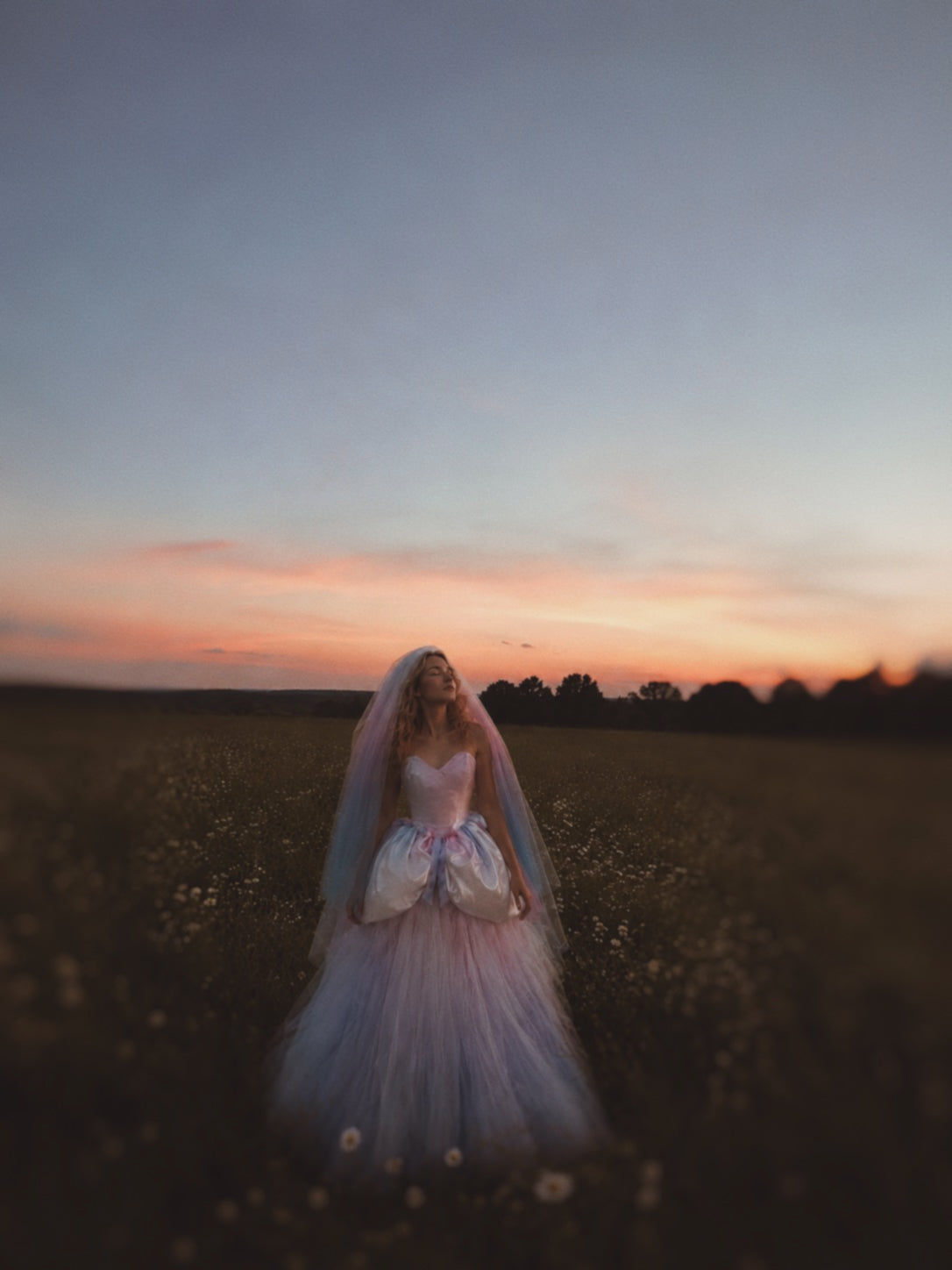 Bride wearing the Moondreamer rainbow wedding dress and ombré veil standing in a wildflower meadow at sunset, featuring a watercolor satin corset bodice and pastel fairytale ball gown skirt.